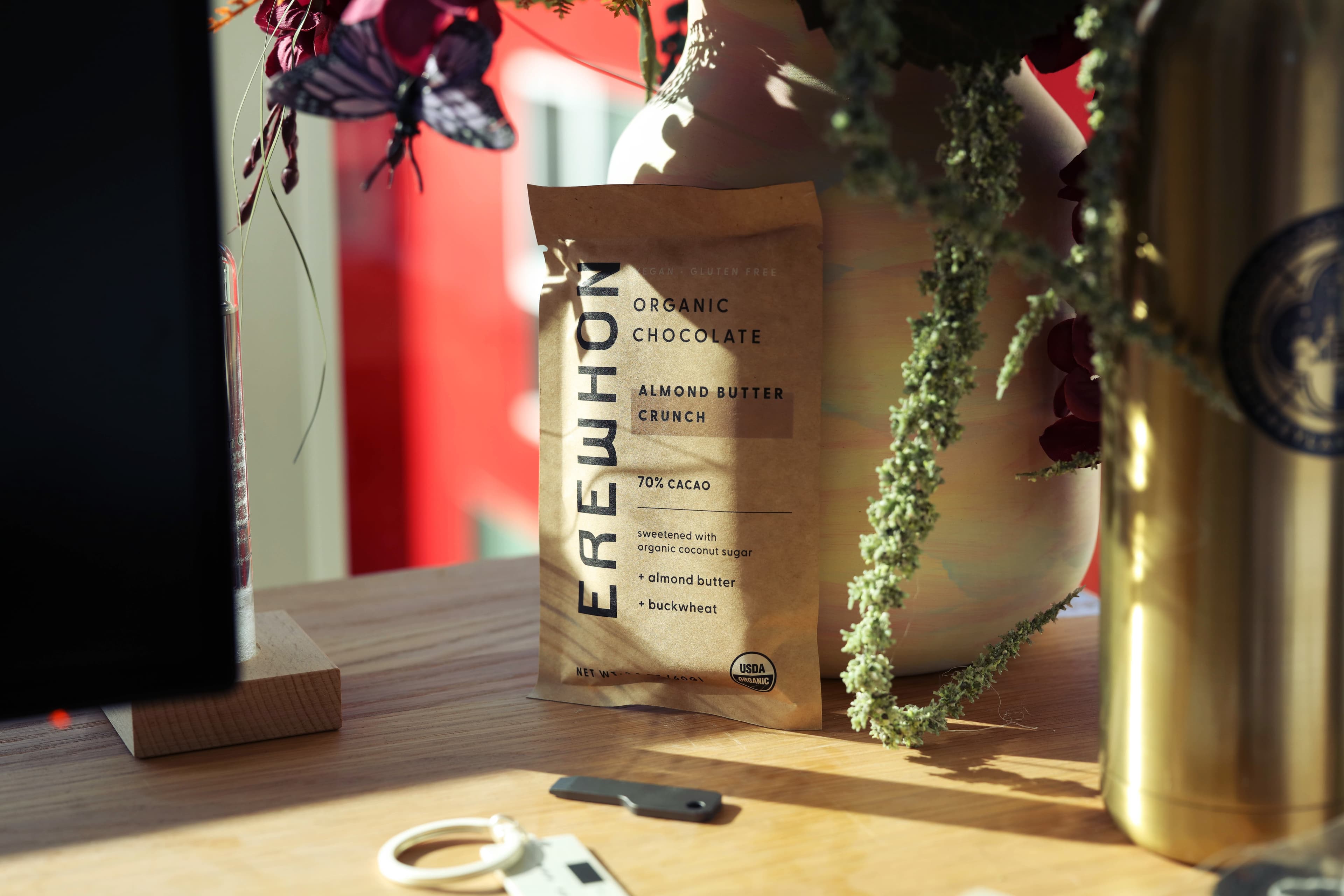Almond Butter Crunch bar made by Unknown for Erewhon in Canada pictured on a messy desk leaning against a flower vase in the sun