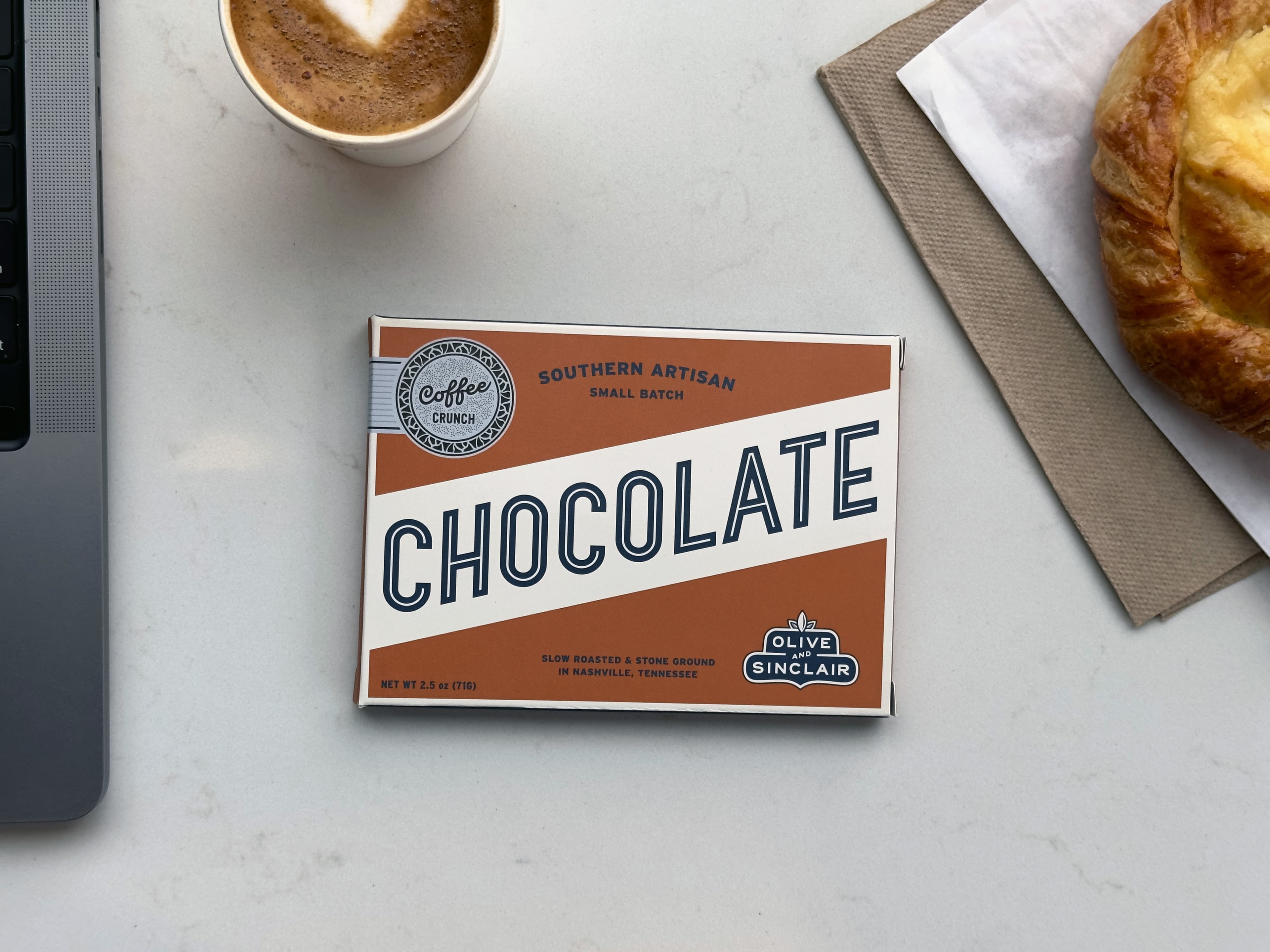 Coffee Crunch bar from Olive & Sinclair Chocolate Co on a marble table with a laptop and coffee cup