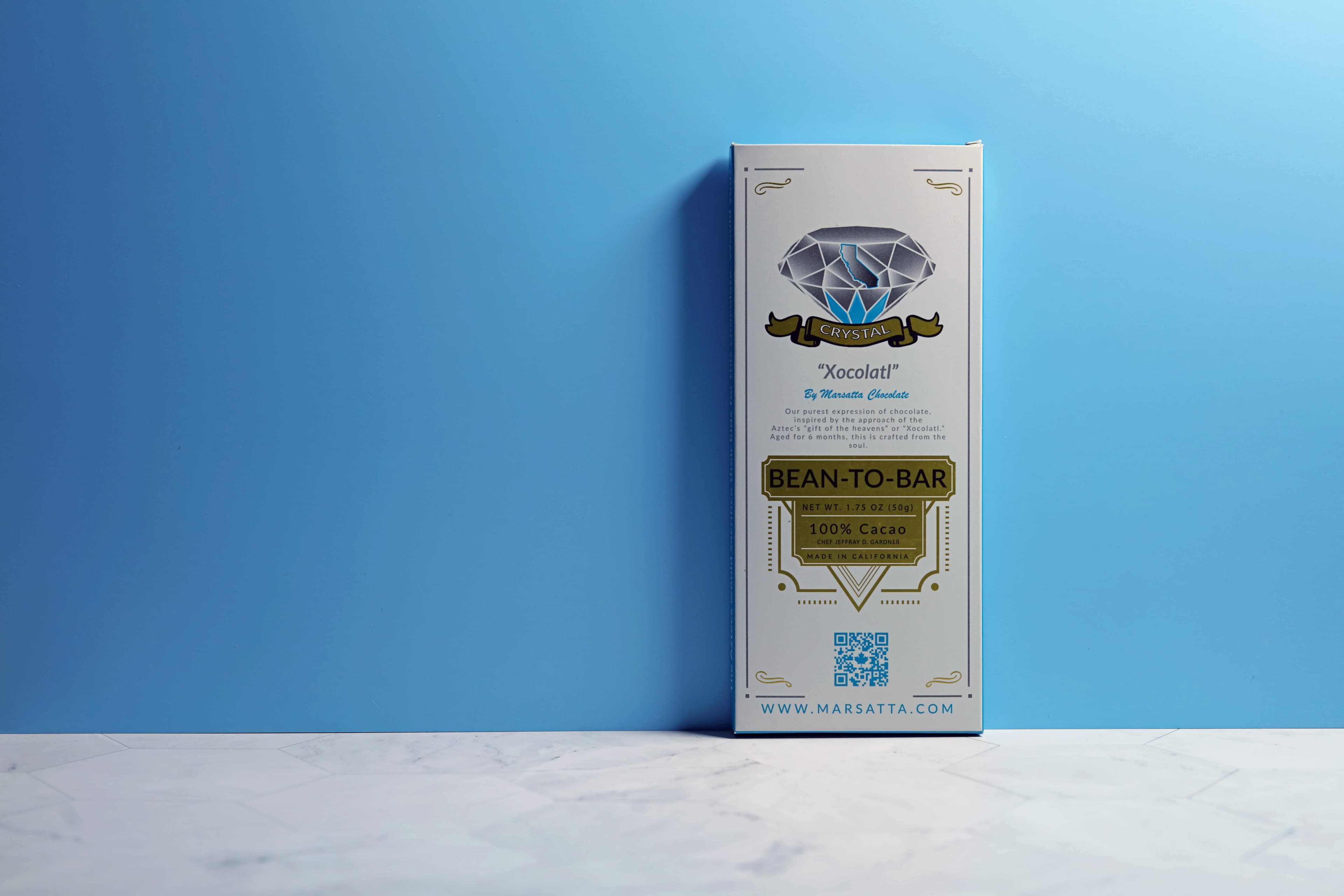 Crystal Label 100% Bean-To-Bar bar made by Marsatta Chocolate in the United States stood up on a marble table with a blue wall behind